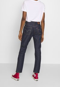 Jeans de mezclilla oscuros de cintura alta con bolsillos traseros curvados, etiqueta de cuero marrón y costuras doradas en contraste, combinados con zapatillas altas rojas.