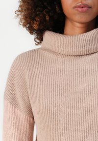 Harmaa turtleneck-neule, jossa on ribbikuviointi ja puoli hihat. Olkapäällä vaaleanpunainen yksityiskohta. Pehmeä, neulottu kangas.