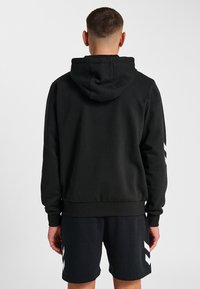 Zwarte hoodie met een textuurafwerking, voorzien van ribben bij de mouwen en de onderrand. Opvallend wit chevronpatroon op de mouwen, gestyled met zwarte shorts.