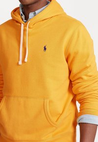 Gul hoodie som är gjord av mjukt tyg. Har en framficka, dragsko i huvan och en lila logotyp på bröstet. Ärmarna är rullade över en blå tröja.