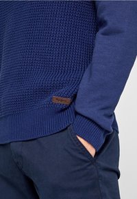 Pull en bleu marine avec tricot texturé sur la partie supérieure et tissu lisse sur les manches. Équipé d'une discrète étiquette de logo marron en bas.