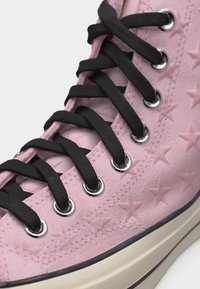 Κοντινή λήψη ροζ σουέντ αθλητικού με αστέρια, μαύρα κορδόνια περασμένα από ασημί κρίκους και χοντρή μπεζ σόλα.