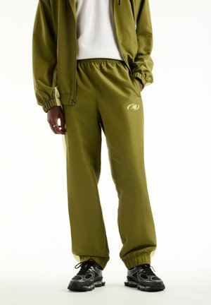 Personne portant un pantalon de survêtement vert olive et une veste assortie avec des chaussures de sport noires, debout devant un fond clair.