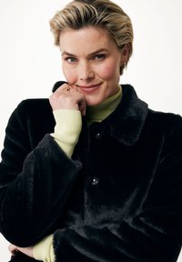 Svart fuskpälsjacka med krage, med knappstängning och långa ärmar. Bärs över en ljusgrön polotröja. Slät textur.