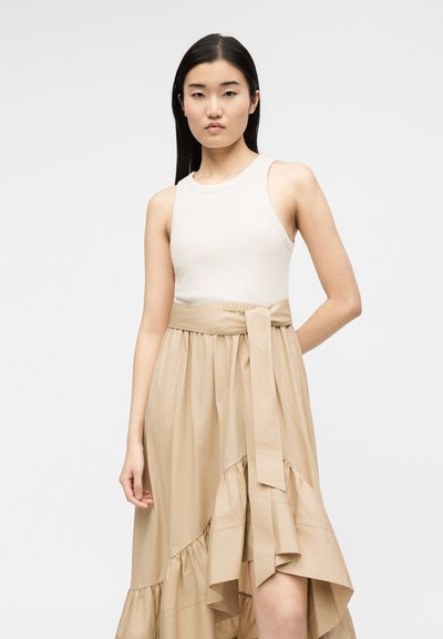 Νεαρή γυναίκα με μακριά σκούρα μαλλιά, που φοράει λευκό αμάνικο τοπ και μπεζ ψηλόμεση φούστα με δεμένη ζώνη, στέκεται μπροστά σε απλό φόντο.