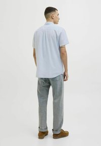 Camisa de manga corta de color azul claro con un ajuste relajado, combinada con jeans de color gris claro. Modelo lleva botas marrones hasta el tobillo. Vista trasera, diseño simple.