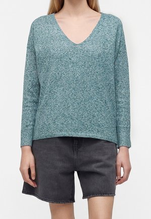 Personne portant un pull à col en V bleu sarcelle et un short en jean noir, debout devant un fond blanc uni.