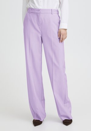 Donna che indossa pantaloni lavanda a vita alta e gamba larga, camicia bianca abbottonata e scarpe scure con punta, in piedi su sfondo bianco.