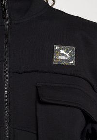 Veste noire zippée avec un petit logo carré multicolore Puma sur le haut de la poitrine et une grande poche à rabat sous le logo.