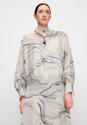 Frau trägt eine transparente Langarmbluse und einen passenden Rock mit abstrakten, wirbelnden Linienmustern in gedämpften Tönen.