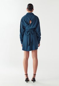 Zelena obleka s kragnico, dolgimi rokavi in pasom na zavezovanje. Ima detajl v obliki ključavnice na hrbtni strani ter gladko, teksturirano tkanino.