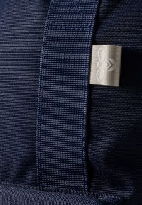Navy stoffen tas close-up, met een textuurpatroon, verstevigde riem en een zilveren logo plaat met reliëf ontwerp.