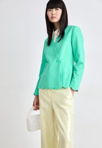Teal blazer met een gegolfde zoom, sluiting met één knoop, gecombineerd met glanzende gele broeken. Model houdt een kleine parelhandtas vast.