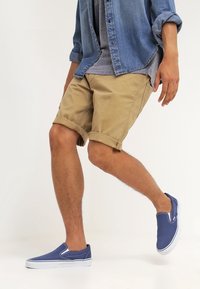 Des chaussures slip-on en toile bleue avec des semelles en caoutchouc blanches, associées à un short beige retroussé et à un t-shirt gris, ainsi qu'une chemise en denim portée ouverte.