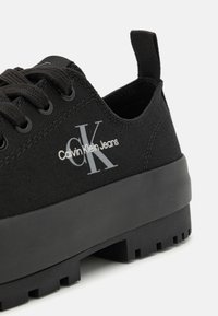 Zapatillas de lona negras con suela de goma plana y diseño texturizado. Presentan un talón elevado, cordones en la parte delantera y un logo en impresión gris.