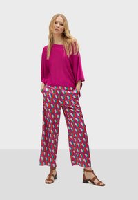 Un haut surdimensionné fuchsia associé à un pantalon large à motifs dans des formes roses, bleues et rouges. Des sandales brunes à lanières complètent le look.