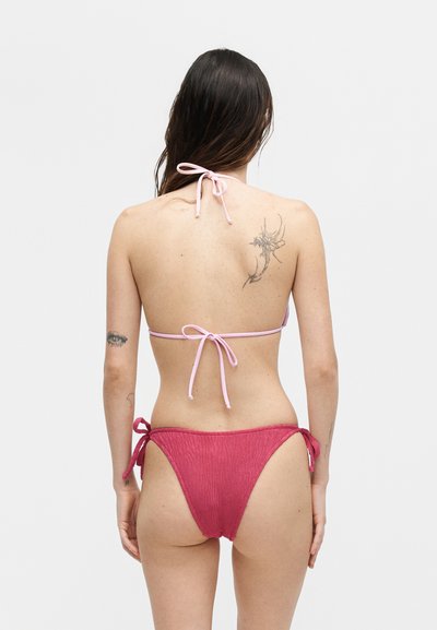 Γυναίκα που φοράει ροζ μπικίνι με δεμένες τιράντες, δείχνοντας την πίσω όψη με τατουάζ στο πάνω μέρος της πλάτης και στο αριστερό χέρι, μπροστά σε φωτεινό απλό φόντο.