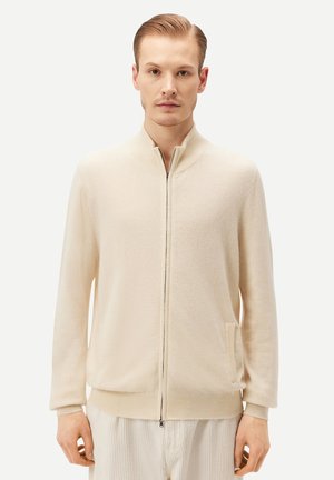 Mann, der einen cremefarbenen Pullover mit Reißverschluss trägt, mit gerippten Bündchen und Saum, vor einem schlichten hellen Hintergrund steht.