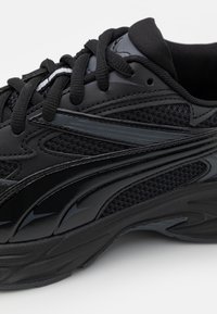 Black athletic sneaker featuring a mesh upper with glossy accents, rounded shape, textured sole, and lace-up closure.
