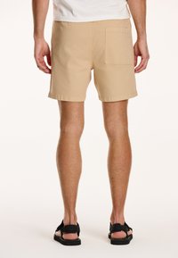 Shorts bej din bumbac, cu un singur buzunar la spate, tiv cusut și o croială relaxată. Purtați cu sandale negre.