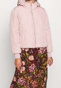 Svetle růžová péřová bunda na zip s kapucí a bočními kapsami. Má prošívanou strukturu a elastické manžety. Nositelná přes květinovou sukni.