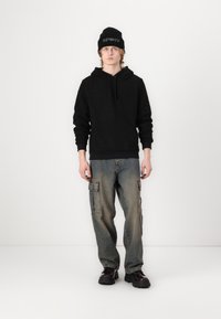 Czarny pluszowy hoodie z kieszenią z przodu, w połączeniu z jasnoniebieskimi spodniami cargo z bocznymi kieszeniami oraz ciemnymi, masywnymi butami. Czarna czapka beanie z napisem.