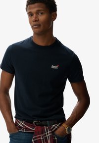 Camiseta de algodón azul marino con cuello redondo, que presenta un pequeño logotipo bordado. Una camisa de cuadros roja y negra atada a la cintura.