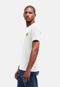 Weißes Baumwoll-T-Shirt mit kurzen Ärmeln, versehen mit einem kleinen gelben Zitronenmotiv, das auf der linken Brust gestickt ist. Dazu werden Jeans aus Denim getragen.