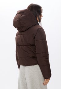 Brązowa kurtka puchowa z kapturem, z pikowanym wzorem i skróconym krojem. Materiał wydaje się gładki i teksturowany.