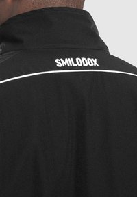Czarna sportowa kurtka z wysokim kołnierzem, z białymi przeszyciami i haftowanym na plecach napisem "SMILODOX". Gładka faktura.