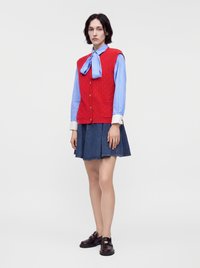 Femme debout portant un chemisier bleu avec un grand nœud, un gilet rouge boutonné, une jupe plissée bleu foncé, et des mocassins marron foncé sur fond blanc.