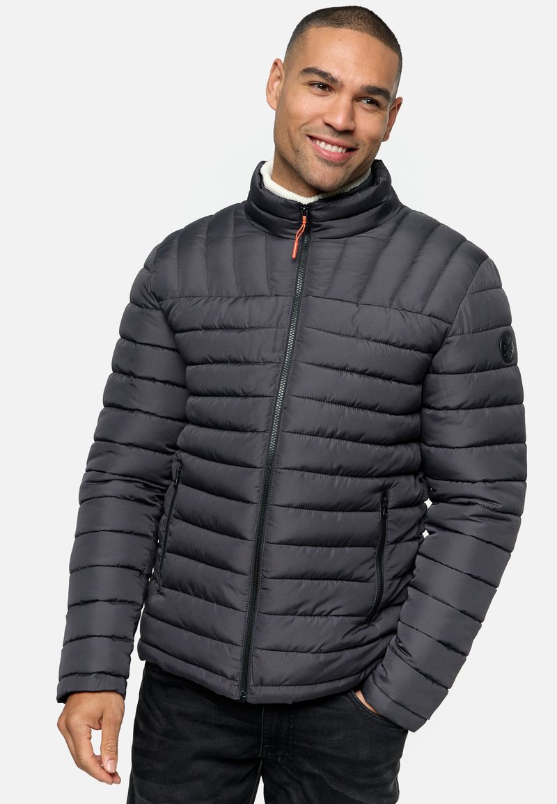 Chaqueta de plumas en gris oscuro, con textura acolchada y cuello alto. Presenta un cierre de cremallera, bolsillos laterales y un logo en la manga.