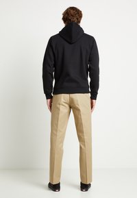 Felpa nera con orlo e polsini a coste, abbinata a pantaloni beige. La felpa presenta un cappuccio con cordino e l'outfit è completato da scarpe nere.