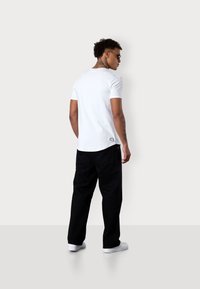 Weißes T-Shirt mit kurzen Ärmeln und abgerundetem Saum, getragen mit schwarzen Hosen und weißen Sneakers; hat subtile Branding am hinteren Saum.