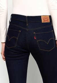 Mørkeblå denimjeans med gule syninger, prydet med to baglommer og et lædermærke med brandlogo ved taljen.
