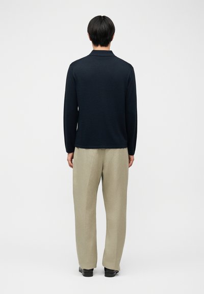 Άνδρας που στέκεται ανάποδα, φοράει μαύρο πουκάμισο με μακριά μανίκια, μπεζ παντελόνι και μαύρα παπούτσια, μπροστά σε λευκό απλό φόντο.