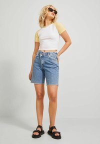 Camiseta corta blanca con mangas amarillas, combinada con pantalones cortos de mezclilla azules con dobladillos desflecados. La modelo lleva sandalias negras y gafas de sol.