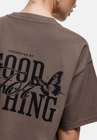 Bruine oversized t-shirt met een zwarte grafische print met tekst en een vlinder op de achterkant. Katoenen materiaal, ontspannen pasvorm, ronde hals.