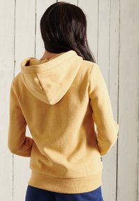Sudadera con capucha amarilla de textura suave, con puños y cinturilla de canalé. Presenta una capucha y un bolsillo tipo canguro. Mostrada desde la parte posterior.