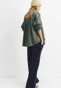 Überprüfte grüne Übergröße-Hemd mit hochgerollten Ärmeln, kombiniert mit weiten marineblauen Hosen. Eine beigefarbene Strickmütze rundet den Look ab.