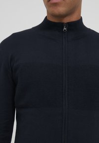 Pull zip bleu marine avec col montant, présentant des sections de tricot texturé et lisse, et une fermeture éclair centrale.