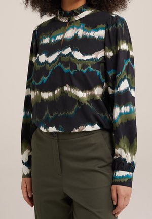 Vrouw draagt een blouse met lange mouwen met groene, blauwe, witte en zwarte abstracte patronen, gecombineerd met een high-waisted olijfgroene broek.
