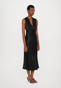 Rochie din satin negru cu decolteu adânc în V, bust ajustat și fustă evazată. Oferă o textură netedă și un luciu subtil. Asortată cu sandale negre cu bretele.