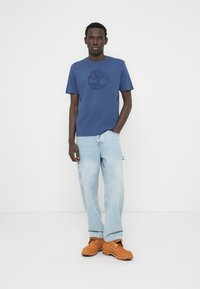 Μπλε μπλουζάκι βαμβακερό με λογότυπο δέντρου, ανοιχτό μπλε τζιν με ίσια γραμμή και καφέ ξύλινα παπούτσια με μαύρη λάστιχη σόλα. Απλή, καθημερινή εμφάνιση.