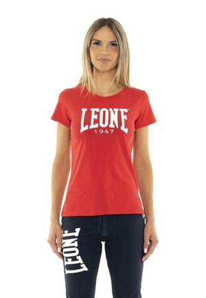 Une femme porte un tee-shirt rouge LEONE 1947 et un pantalon de jogging bleu marine avec un logo blanc, sur un fond blanc uni.