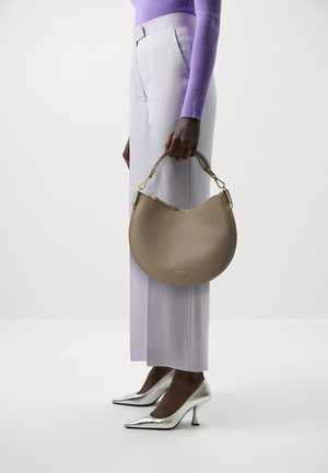 Borsa a tracolla in pelle beige con forma a mezzaluna, dettagli in metallo dorato, tenuta da una persona con pantaloni lavanda e tacchi argento.