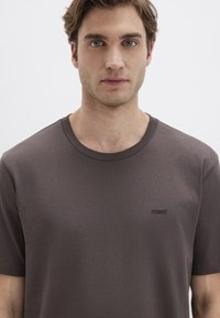 Braunes Rundhals-T-Shirt aus glattem Stoff, mit kurzen Ärmeln und einem kleinen schwarzen Logo auf der linken Brust.