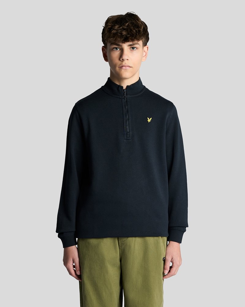 Νεαρός άνδρας με κοντά σγουρά μαλλιά, φοράει μαύρο πουλόβερ με φερμουάρ έως το στήθος και ελιάς πράσινου χρώματος παντελόνια, στέκεται μπροστά σε ένα απλό γκρι φόντο.