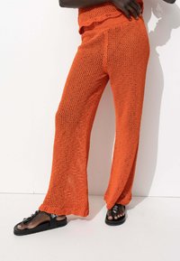 Pantalons large en tricot orange avec un motif de tissage lâche et ouvert ; taille et ourlet festonnés ; associés à des sandales noires ornées.