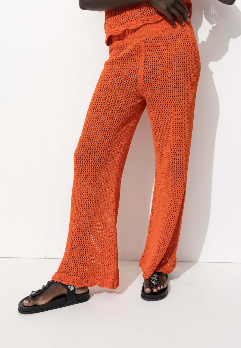 Pantalons large en tricot orange avec un motif de tissage lâche et ouvert ; taille et ourlet festonnés ; associés à des sandales noires ornées.